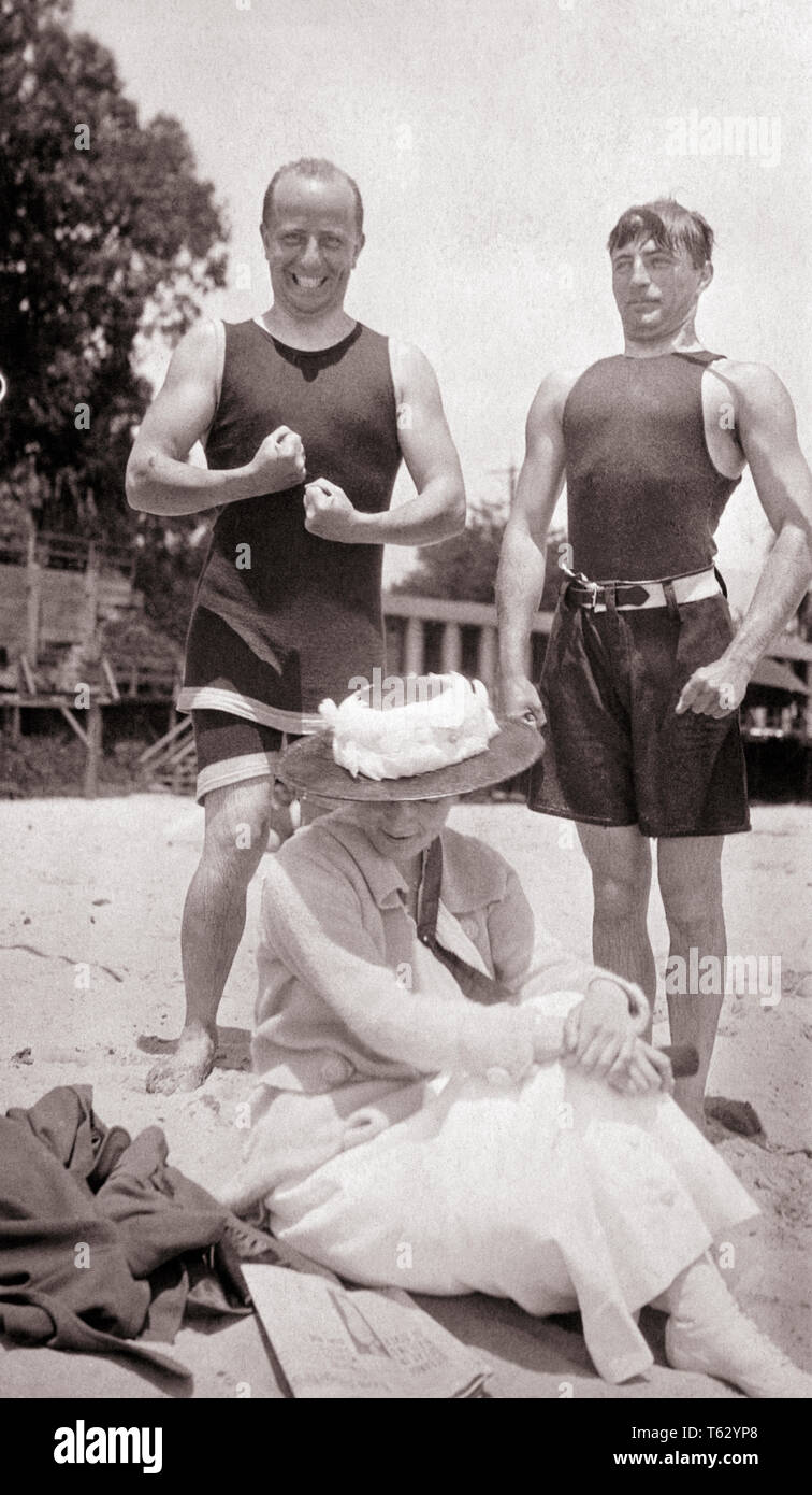 1910s TWO ANNOYING MEN SUITORS IN BATHING COSTUMES FLEXING THEIR MUSCLES AT  EMBARRASSED WOMAN HIDING UNDER HAT SITTING ON BEACH - o3870 HAR001 HARS  STYLE BATHING YOUNG ADULT COMIC COMPETITION SUITS PLEASED