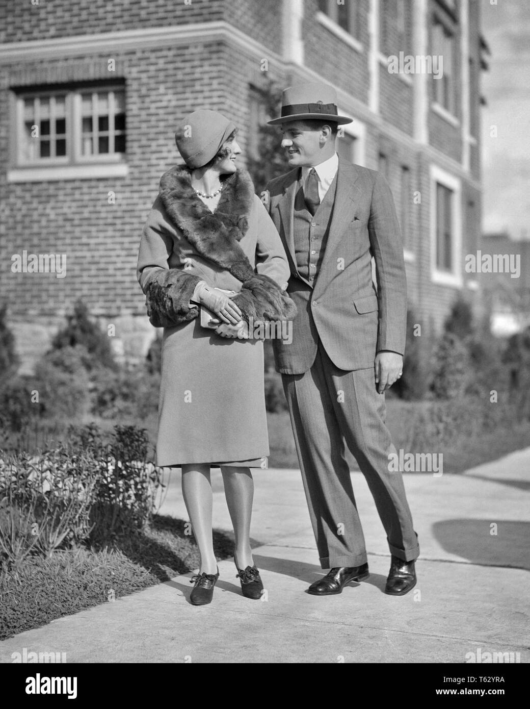 1920s SMILING COUPLE MAN WOMAN WALKING ARM IN ARM WOMAN WEARING COAT WITH FUR TRIM - p2488 HAR001 HARS LIFESTYLE FEMALES MARRIED SPOUSE HUSBANDS HEALTHINESS HOME LIFE COPY SPACE FRIENDSHIP FULL-LENGTH LADIES PERSONS MALES B&W SUIT AND TIE HAPPINESS STYLES CLOCHE TRIM CONNECTION STYLISH COOPERATION FASHIONS MID-ADULT MID-ADULT WOMAN TOGETHERNESS WIVES YOUNG ADULT WOMAN BLACK AND WHITE CAUCASIAN ETHNICITY HAR001 OLD FASHIONED Stock Photo