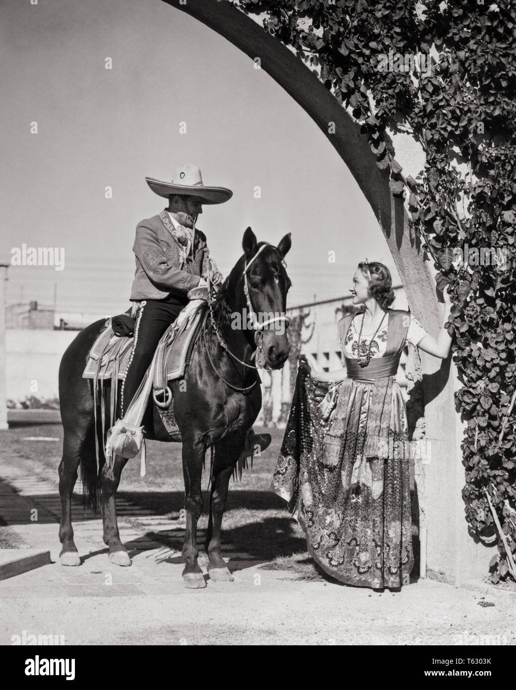 1930s SMILING CAUCASIAN WOMAN IN TRADITIONAL CHINA POBLANA COSTUME TALKING TO MEXICAN MAN HORSEACK WEARING CHARRO COWBOY CLOTHES - r11452 HAR001 HARS LIFESTYLE HISTORY FEMALES RURAL COPY SPACE FRIENDSHIP FULL-LENGTH LADIES PERSONS TRADITIONAL MALES TRANSPORTATION MEXICAN B&W NORTH AMERICA MEXICO ADVENTURE STYLES LATIN AMERICAN STYLISH FASHIONS MID-ADULT MID-ADULT MAN YOUNG ADULT WOMAN BLACK AND WHITE CAUCASIAN ETHNICITY CHARRO CHINA POBLANA HAR001 HISPANIC ETHNICITY OLD FASHIONED TOURISM Stock Photo
