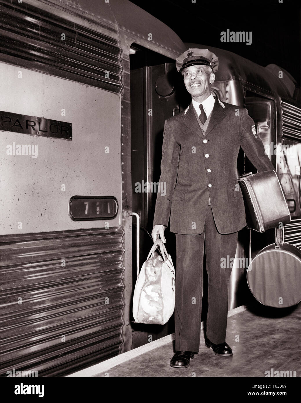 1950s SMILING AFRICAN AMERICAN MAN REDCAP PORTER CARRYING LUGGAGE BY SIDE  OF PASSENGER RAILROAD TRAIN - r190 HAR001 HARS LIFESTYLE JOBS PASSENGER  FULL-LENGTH PERSONS MALES TRANSPORTATION B&W SKILL OCCUPATION PLATFORM  SKILLS CHEERFUL