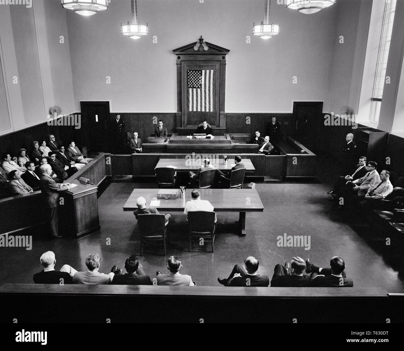 1960s COURTROOM AMERICAN FLAG HANGING BEHIND JUDGE JURY ON RIGHT LAWYER AT LECTERN SPEAKING - s12370 HAR001 HARS LADIES PERSONS INSPIRATION UNITED STATES OF AMERICA MALES B&W GOVERNMENT TABLES FREEDOM HEAD AND SHOULDERS HIGH ANGLE DISCOVERY STRATEGY JUDICIAL LAWYERS POWERFUL JUDGMENT RIGHTS ATTORNEYS AUTHORITY POLITICS RIGHT CONCEPTUAL PROSECUTION ARCHITECTURAL DETAIL ATTORNEY COOPERATION DEFENSE DEMOCRACY IDEAS LECTERN TOGETHERNESS BLACK AND WHITE CAUCASIAN ETHNICITY FREEDOMS HAR001 OLD FASHIONED Stock Photo
