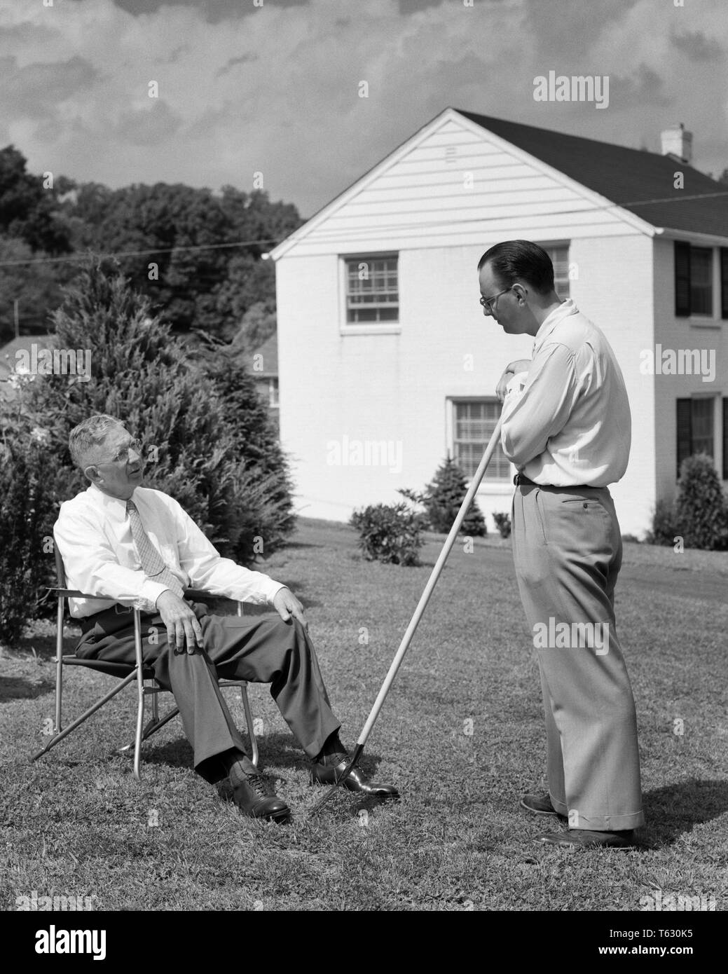 1950s two men in backyard neighbors talking one seated other standing with rake - s5808 HAR001 HARS JUVENILE COMMUNICATION SONS LIFESTYLE ELDER HOUSES RAKE HOME LIFE SEATED COPY SPACE FRIENDSHIP FULL-LENGTH PERSONS RESIDENTIAL MALES BUILDINGS SENIOR MAN SENIOR ADULT FATHERS B&W NEIGHBORS OLDSTERS OLDSTER DADS EXTERIOR FATHER AND SON IN HOMES ELDERS CONNECTION OLD AND YOUNG RESIDENCE STYLISH COOPERATION JUVENILES LAWN CHAIR MID-ADULT MID-ADULT MAN TOGETHERNESS BLACK AND WHITE CAUCASIAN ETHNICITY HAR001 OLD FASHIONED Stock Photo