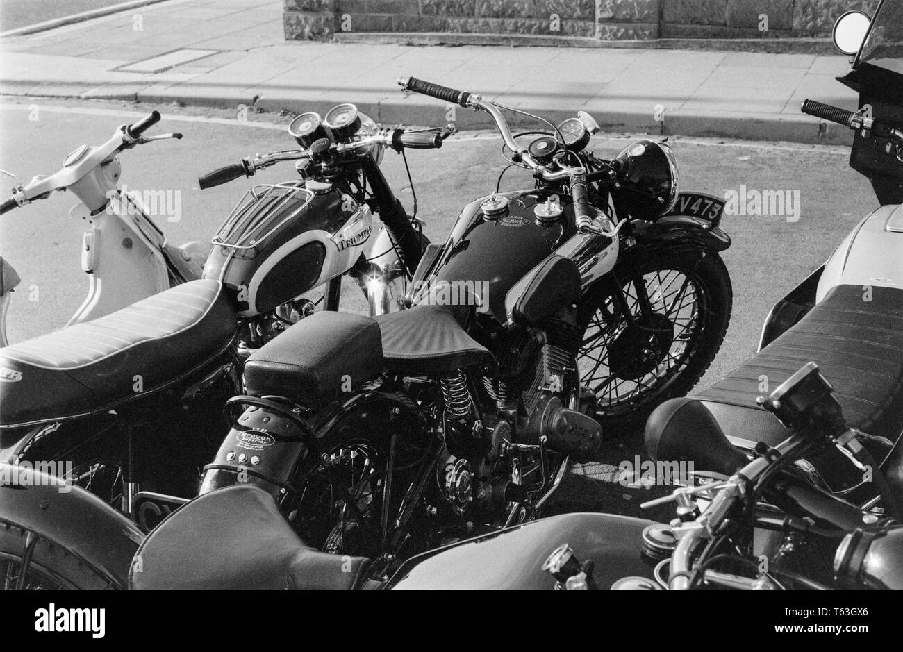 A black and white photograph taken in the 1970s showing details of a Brough Superior SS80 Motorcycle, and a Triumph Motorcycle, both classic British motorbikes. A moped can also be seen. Stock Photo
