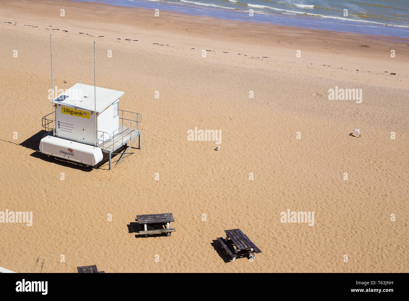 Broadstairs, Kent. UK. A white lifeguard hut on an empty sandy beach in spring on a sunny day. Two empty picnic tables can be seen. Stock Photo
