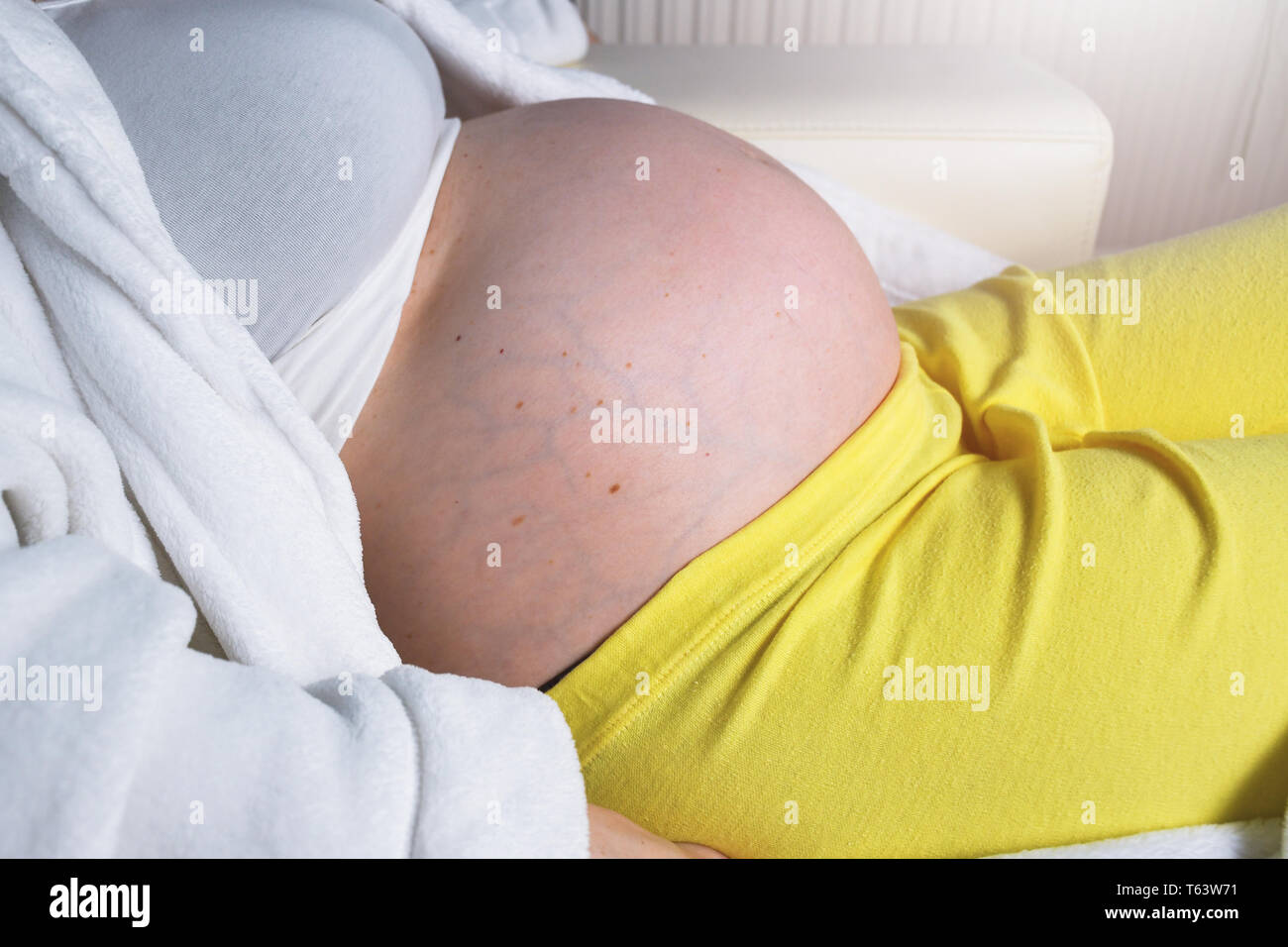 Pregnant woman wearing white bathrobe relaxing and resting at home on a white couch thinking and expecting her newborn baby Stock Photo