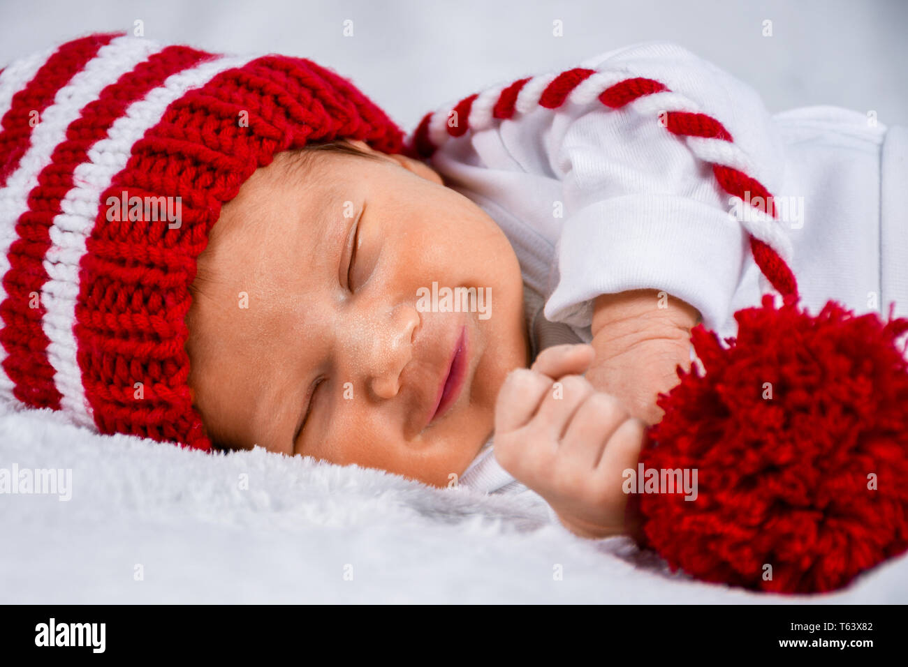 Close up head photo of a cute happy looking adorable newborn baby with red and white striped Christmas theme long tail hat smiling while sleeping Stock Photo