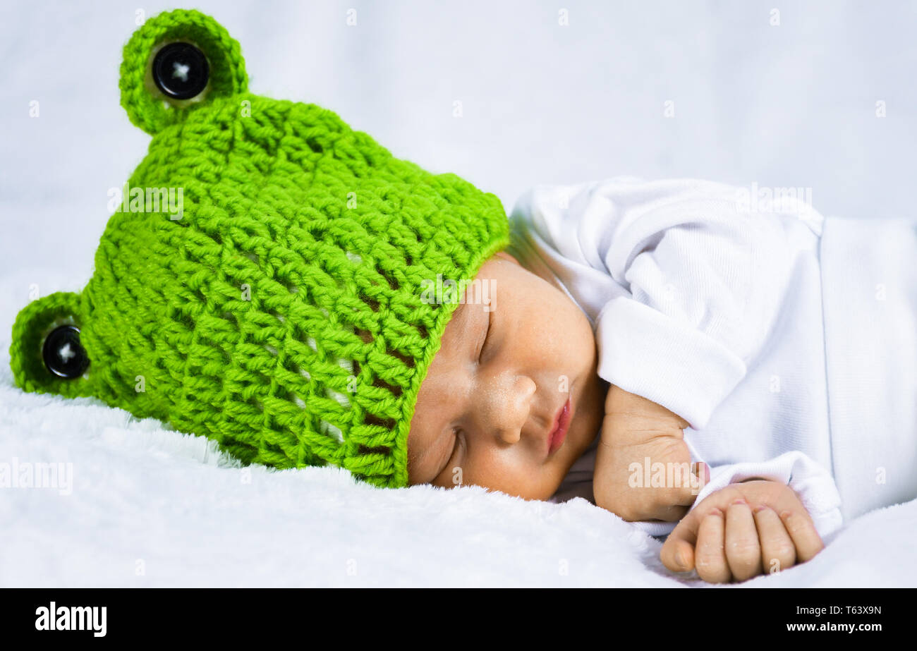 Close up photo of a cute looking adorable newborn baby wearing green frog theme hat with ears peacefully sleeping on a white soft blanket Stock Photo