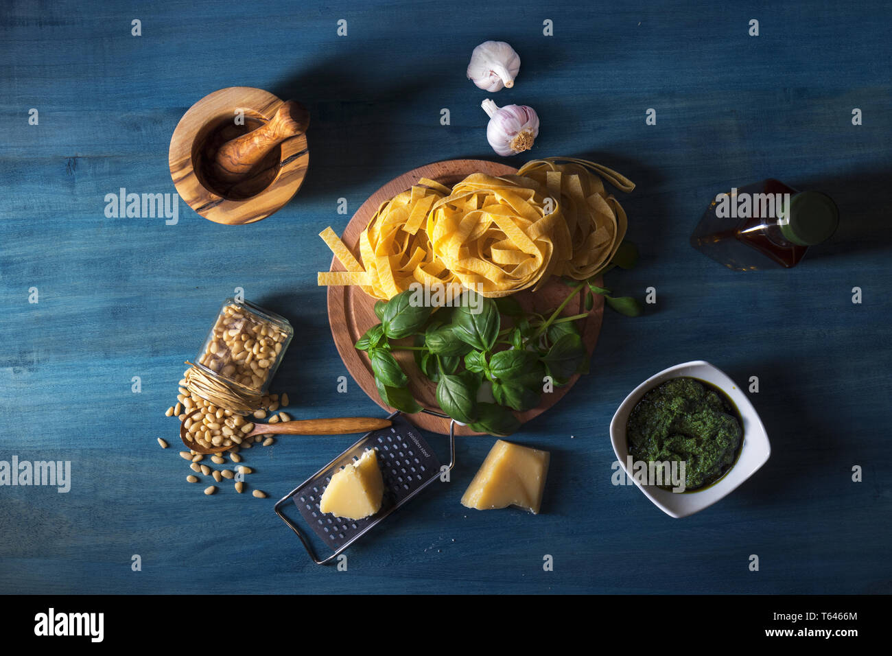 The table with ingredients to produce basil pesto. Mortar, fresh basil herbs, pine nuts, olive oil, parmesan cheese, garlic, bowl with pesto, grater,  Stock Photo