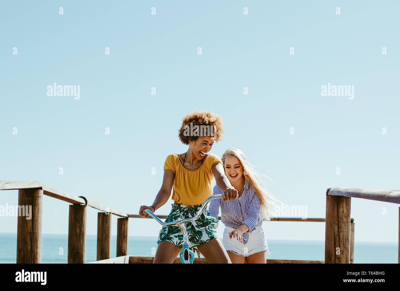 Two women friends playing with a bicycle on the boardwalk along the sea. Woman sitting on the bike with her friends pushing from the back and smiling. Stock Photo