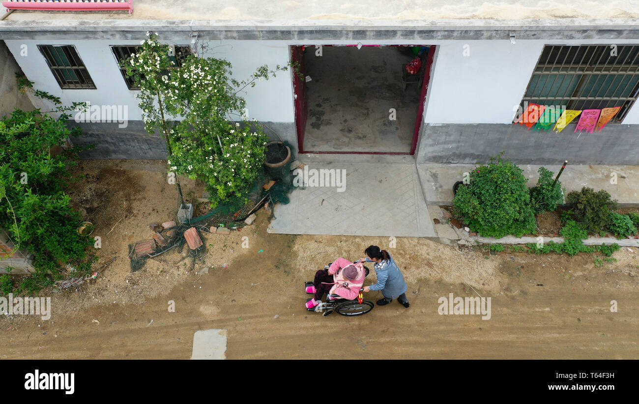 (190429) -- NANJING, April 29, 2019 (Xinhua) -- Aerial photo taken on April 24, 2019 shows Yin Ziyu (R) walking with her mother Liu Jihua on a wheelchair in front of their home at Shanjian Village of Lizhuang Township in Lianyungang City, east China's Jiangsu Province. Yin Ziyu, a 23-year old villager in Shanjian Village, is widely known in the village for her story of taking care of her disabled mother Liu Jihua. Yin's father died of liver cancer when she was four years old. Since then, she and her grandparents took care of her mother Liu living with congenital rickets. At the age of 15, Yin Stock Photo