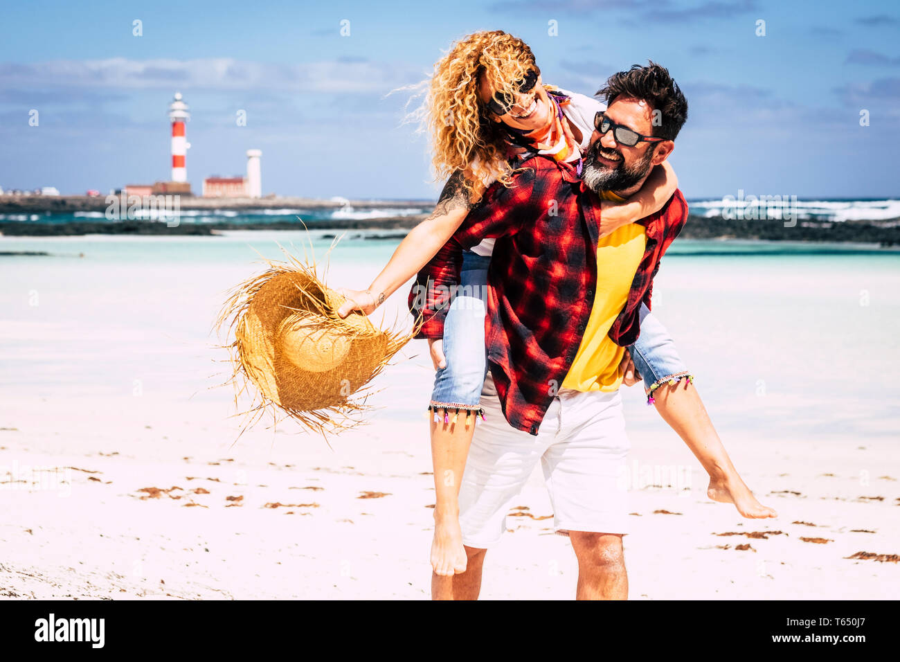 Cheerful people happy joyful couple playing together in love with man carrying woman laughing a lot having fun at the beach in summer holiday vacation Stock Photo
