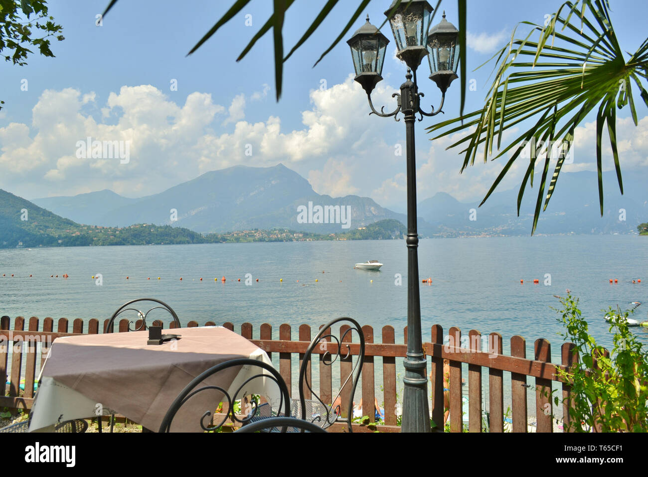 Table with tablecloth, wooden fence, palmtree and streetlamp of Lierna bar on beach with Bellagio peninsula and lake Como with motorboat anchored. Stock Photo