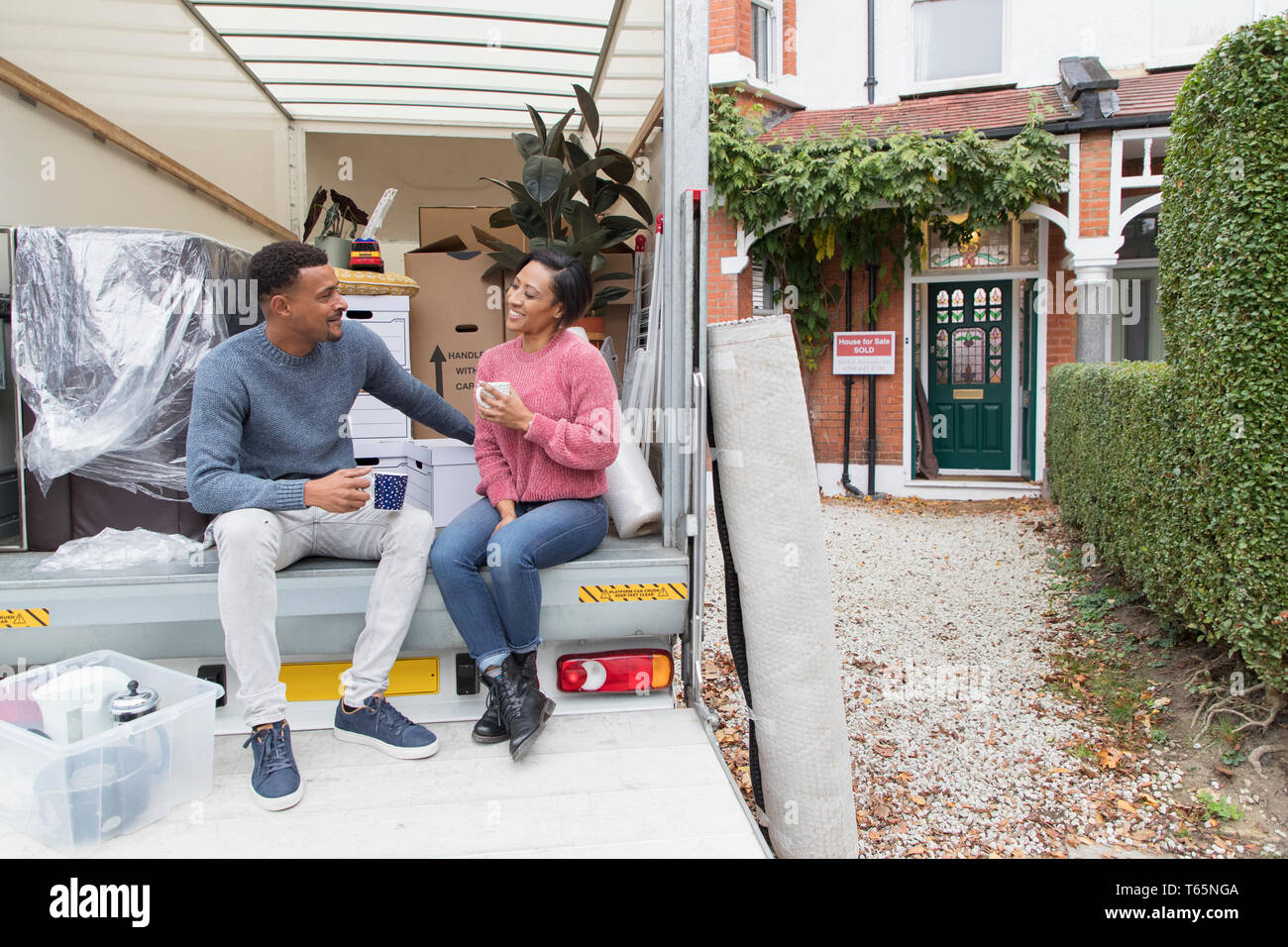 Couple talking and drinking tea, taking a break at back of moving van outside new house Stock Photo