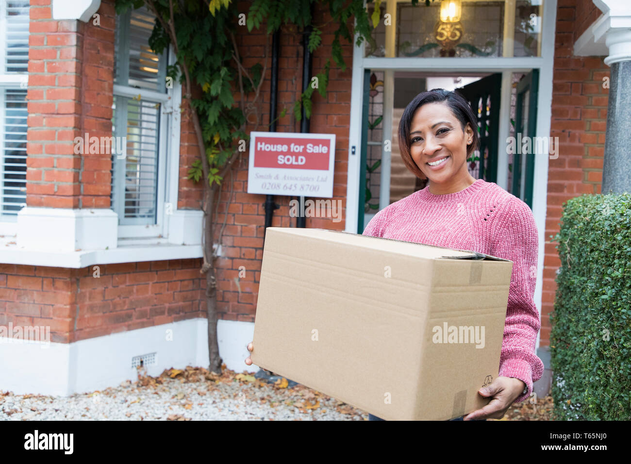 Portrait smiling, confident woman moving into new house, holding cardboard box outside house Stock Photo