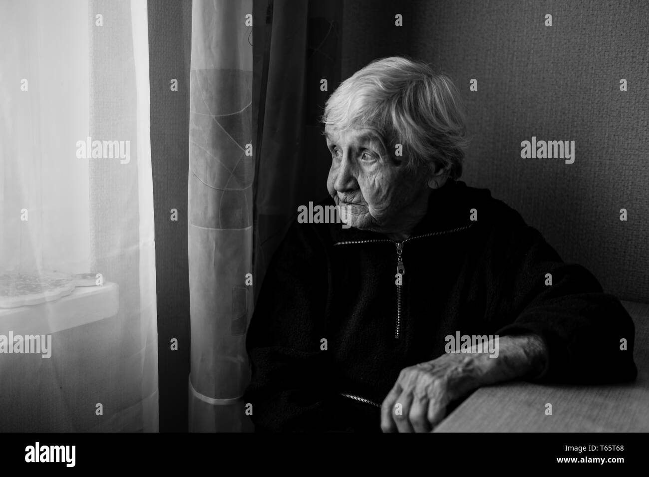 An elderly woman in his house looks out the window longingly. Old lady retired. Black and white photo. Stock Photo