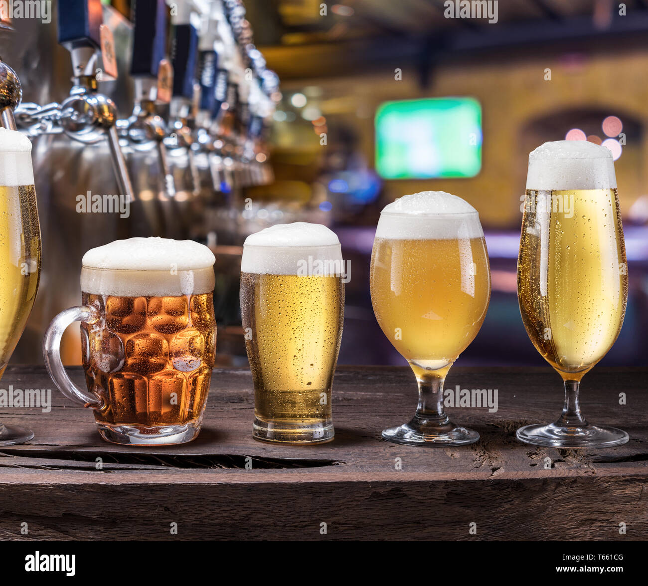 Chilled mugs and glasses of beer on the old wooden table. Pub interior and bar counter with beer taps at the background. Assortment of beer. Stock Photo