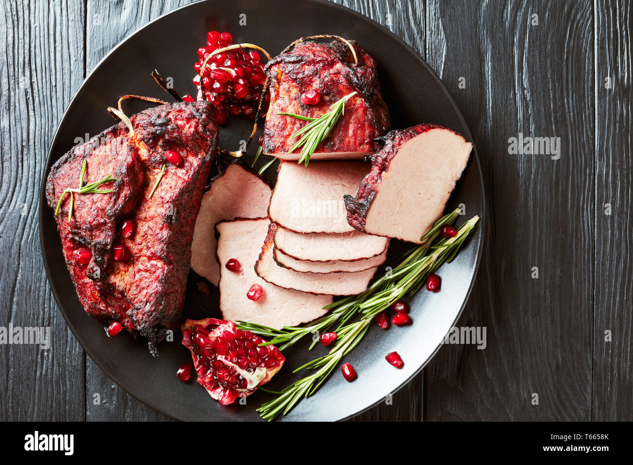 sliced smoked barbecue pork tenderloin on a black platter with fresh garnet seeds and rosemary, on a black wooden table, view from above, flatlay,clos Stock Photo