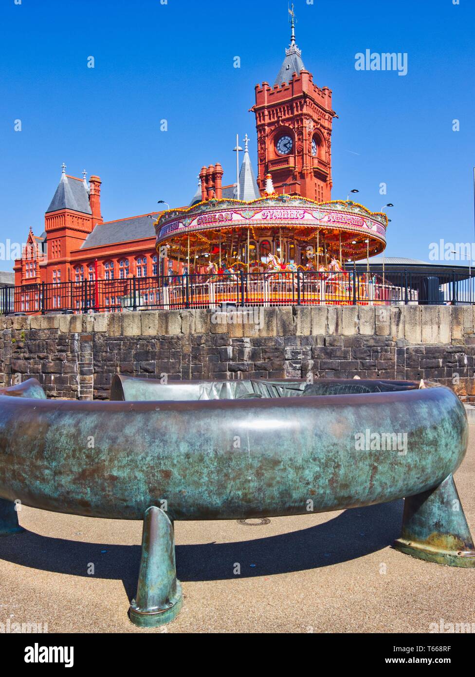 Bronze Celtic Ring sculpture with fairground carousel and the Pierhead building in background, Cardiff Bay, Cardiff, Wales, United Kingdom Stock Photo