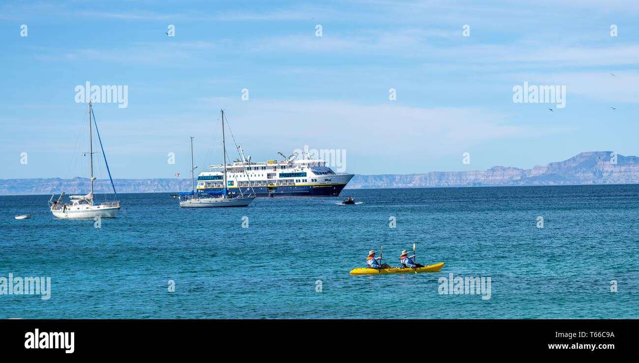 A couple paddle a yellow kayak with sailboats and the National Geographic Venture cruise ship in the background off shore of Isla San Francisco, Baja  Stock Photo
