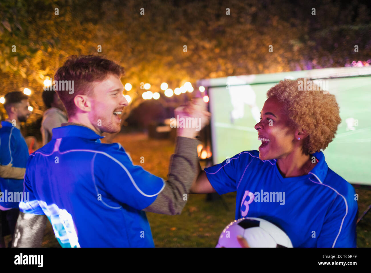 Happy friends cheering, watching soccer match on projection screen in backyard Stock Photo