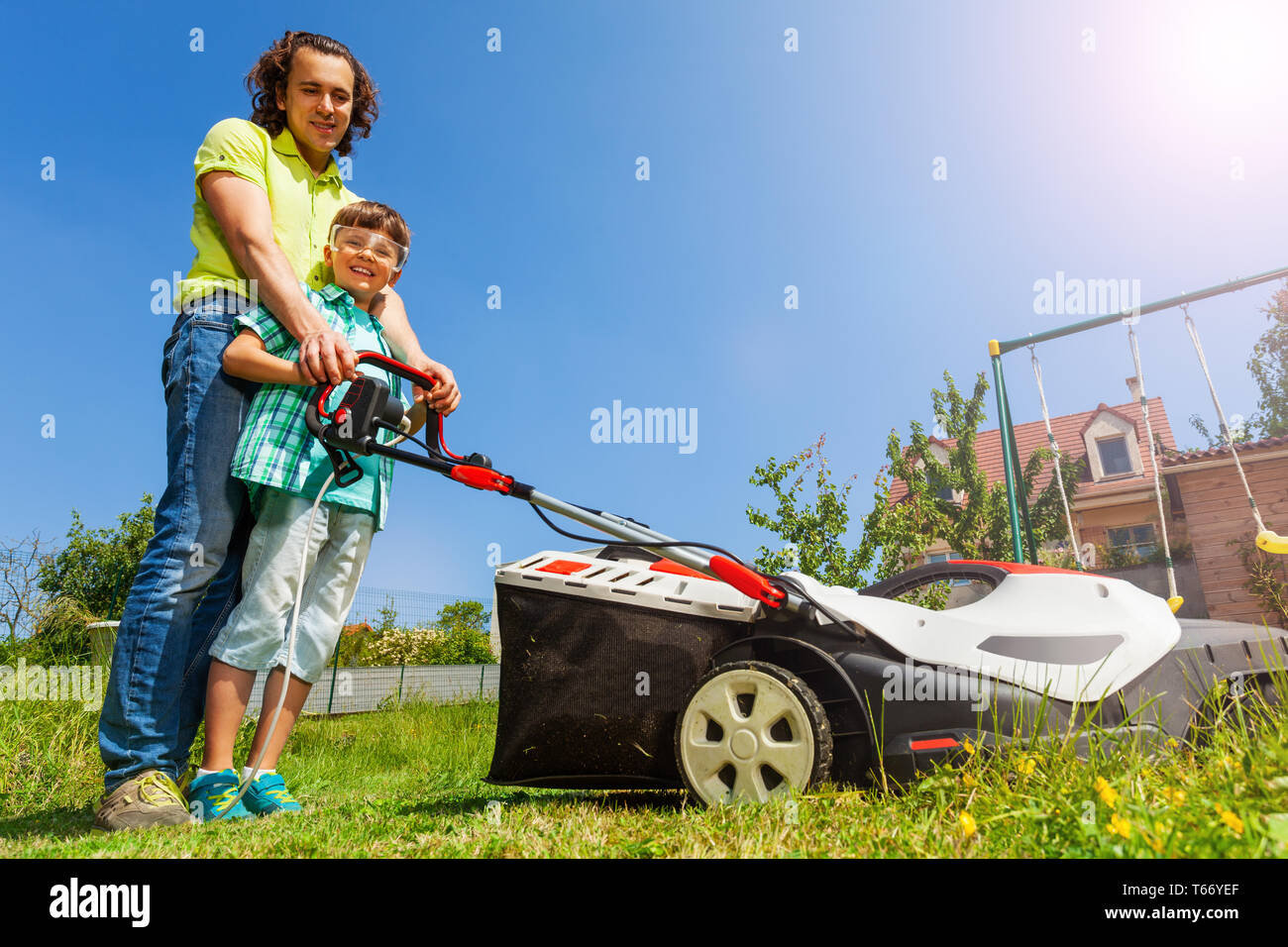 Low angle portrait of young father teaching his son edging the lawn using electricity-powered lawnmower at the backyard Stock Photo