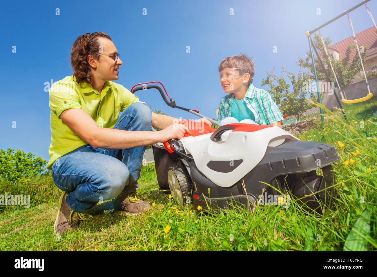 Young father and his son working together at the garden, cutting grass using lawnmower at sunny day Stock Photo