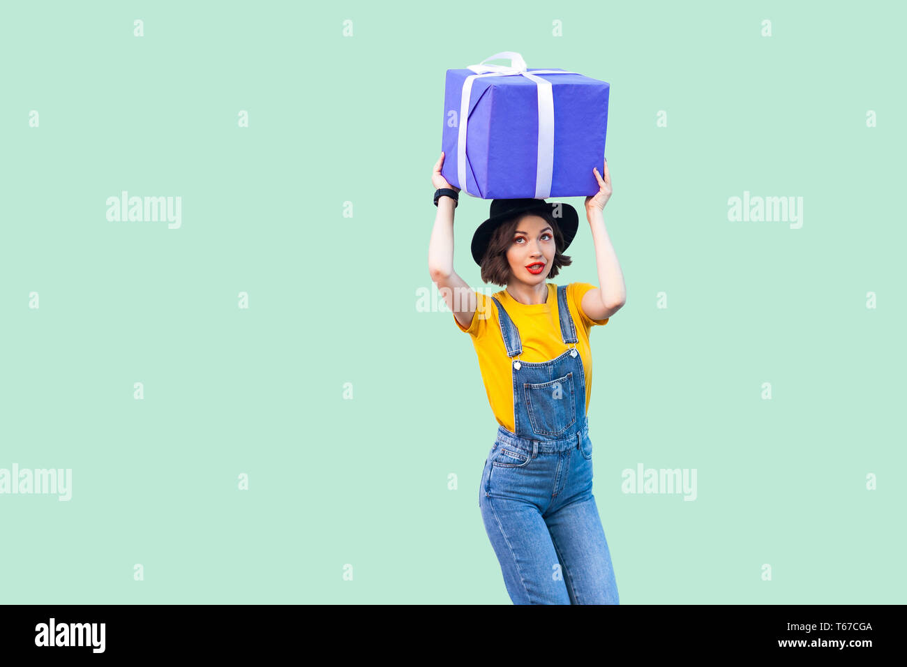 Dreaming positive young girl in hipster wear in denim overalls and black hat standing and holding under head giant big heavy gift box with pondering f Stock Photo