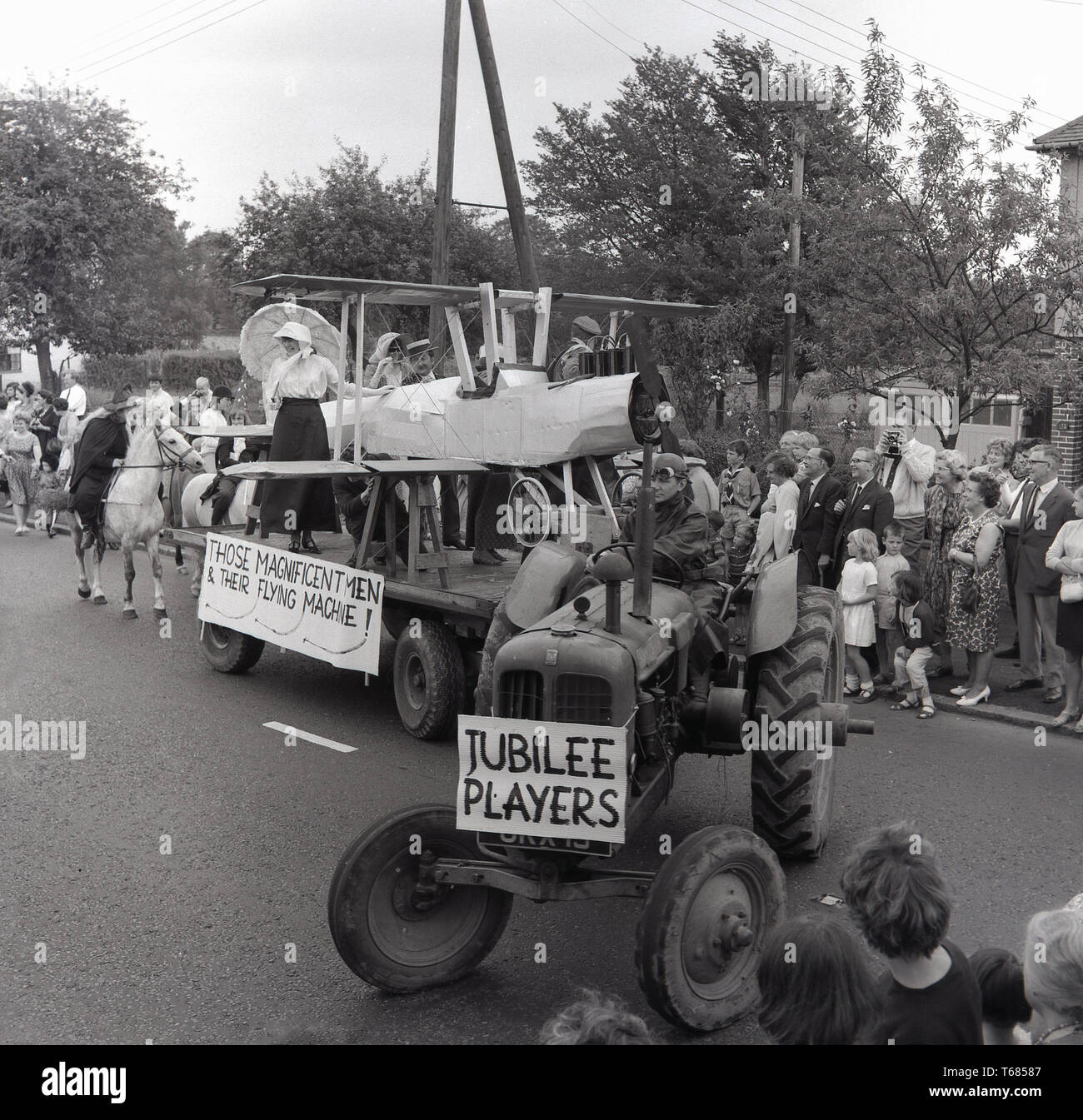 1960s, historical, spectators watch the local village carnival parade go pass, with a tractor pulling a float with a hand-made cardboard bi-plane on, followed by a witch on a horse, Prestwood, Bucks, England, UK. Stock Photo