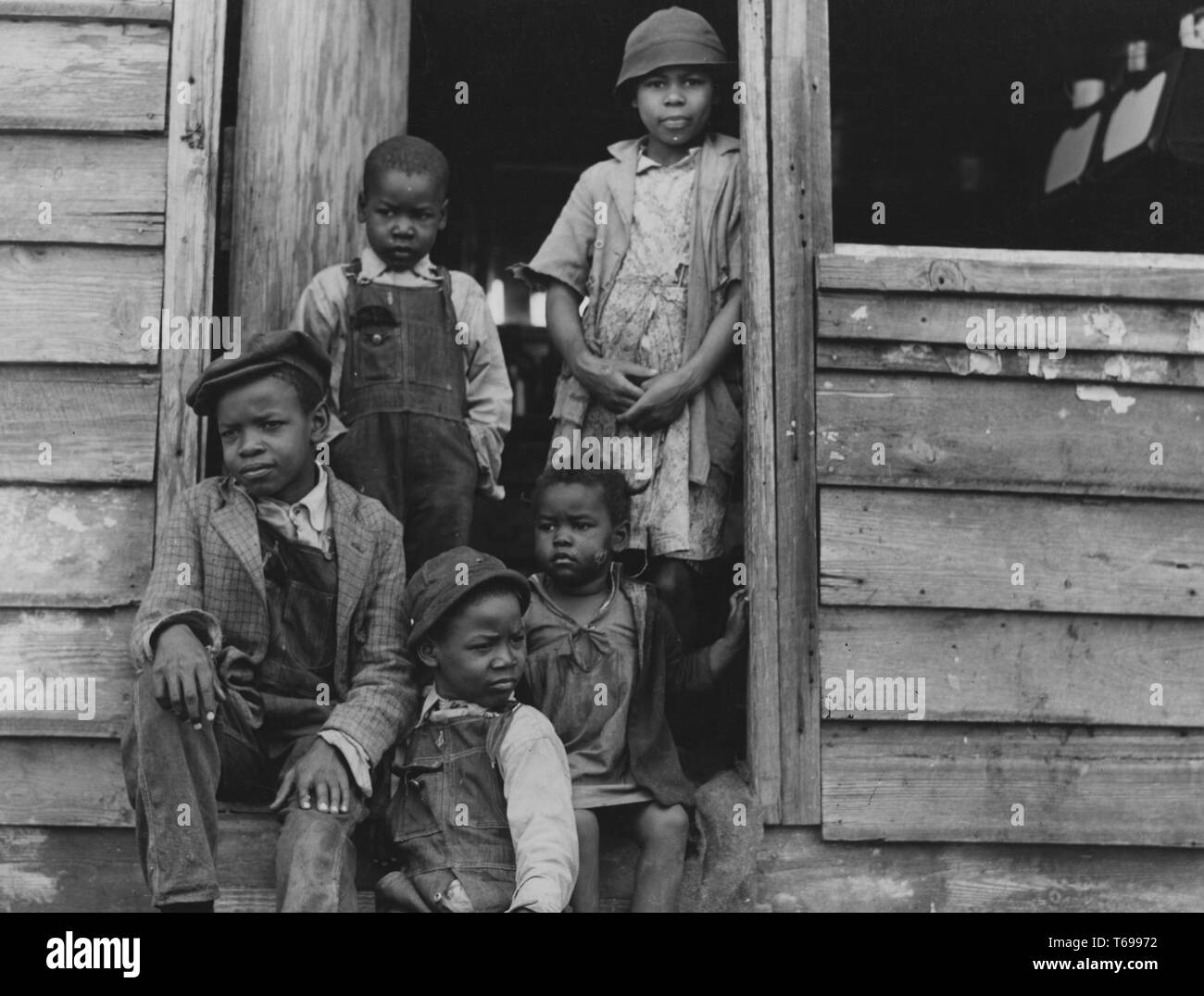 Black and white photograph of three boys and two girls (the oldest girl, wearing a cloche hat and facing the camera) sitting and standing in the doorway to a weathered, wooden building; located in South Carolina, USA; photographed by Jack Delano under the sponsorship of the United States' Farm Security Administration, 1940. From the New York Public Library. () Stock Photo