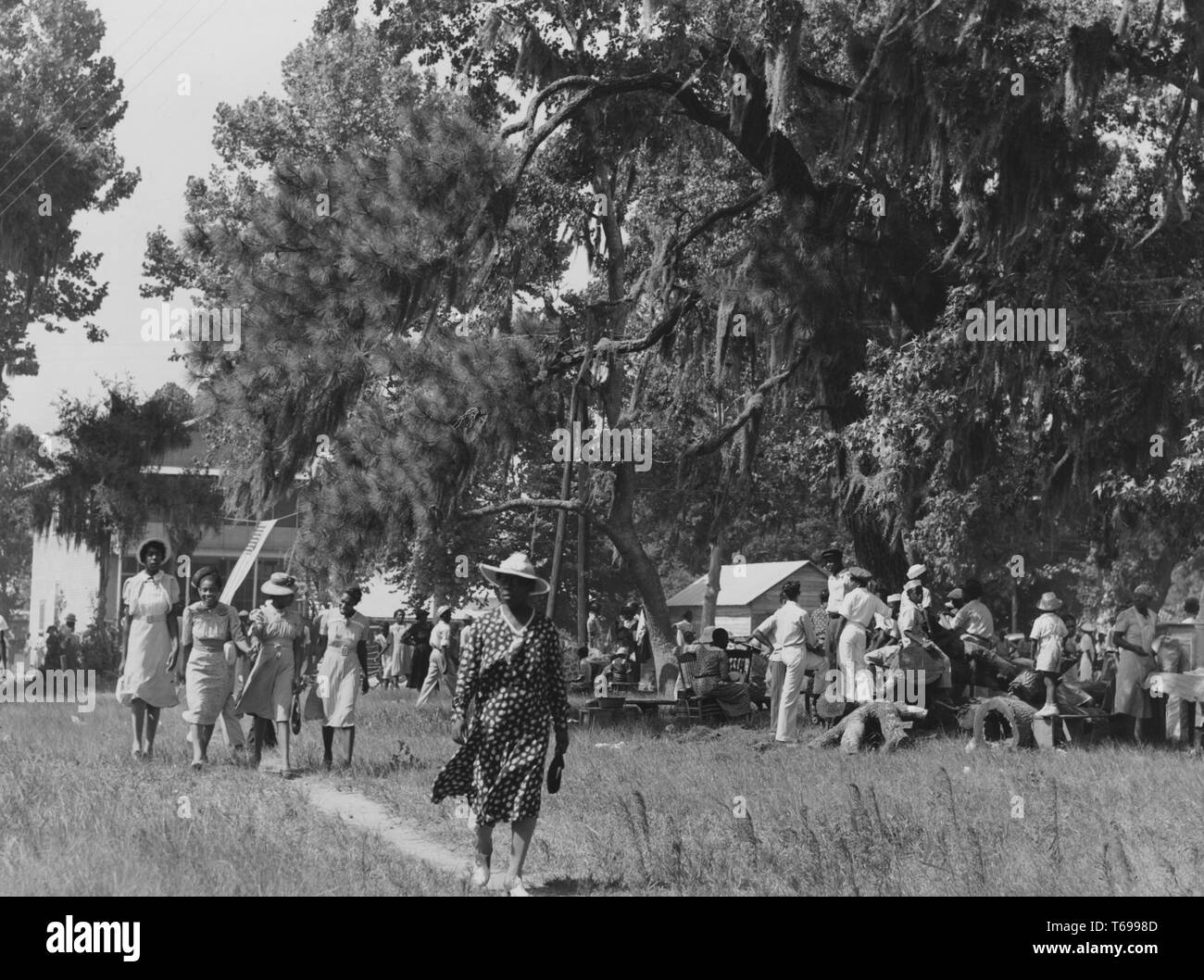 Black and white photograph of a group of African American men and women, enjoying themselves at a picnic or garden party; with a woman, wearing a large hat and a polka-dotted dress, walking toward the camera in the foreground, followed by a row of four young women wearing pale dresses, and with large trees and several buildings visible in the background; located in Beaufort, South Carolina, USA; photographed by Marion Post Wolcott, under the sponsorship of the United States' Farm Security Administration, July, 1939. From the New York Public Library. () Stock Photo