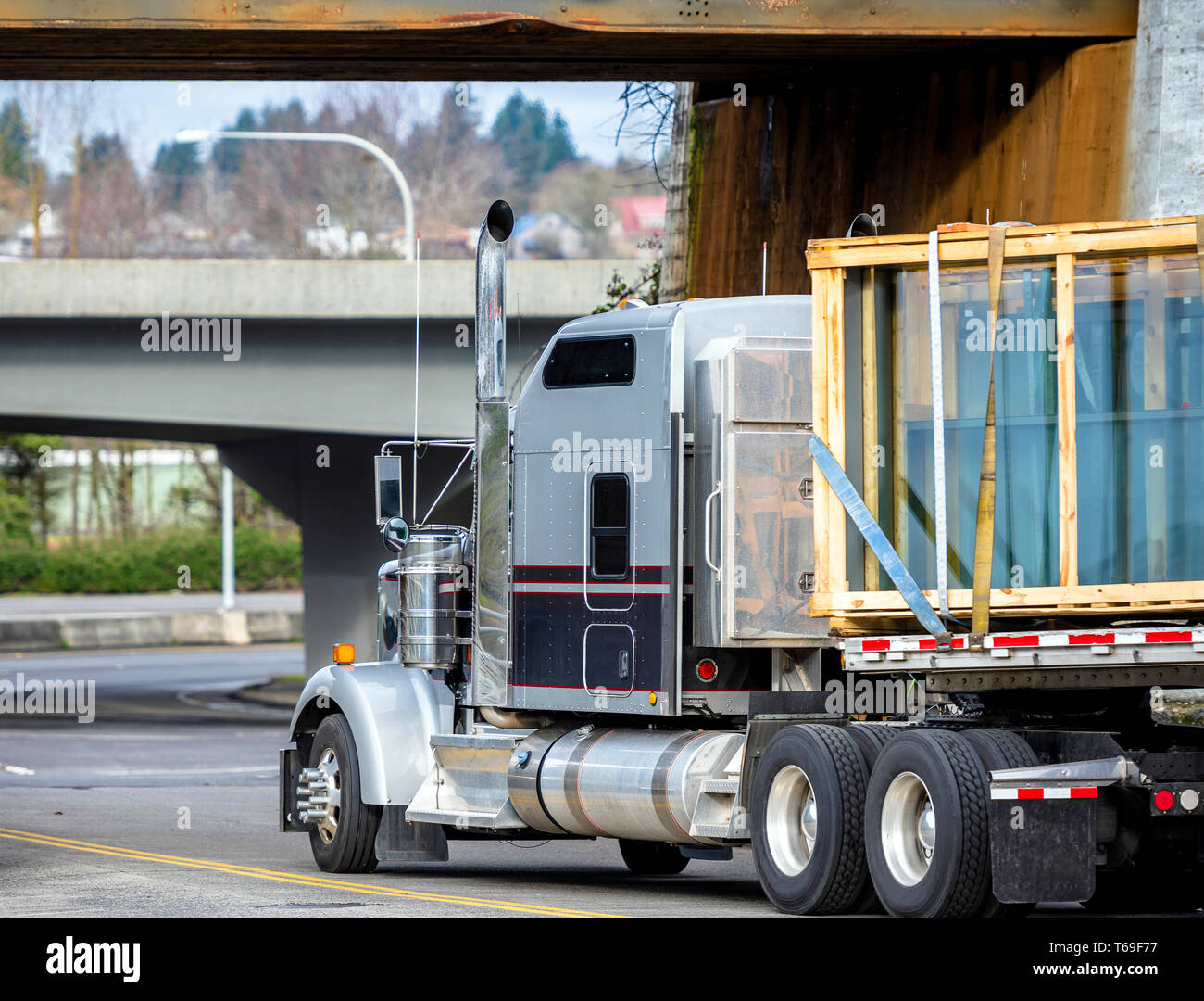 Big rig gray and black classic long haul semi truck transporting windows glass sheets in wood boxes fastened on flat bed semi trailer turning under lo Stock Photo