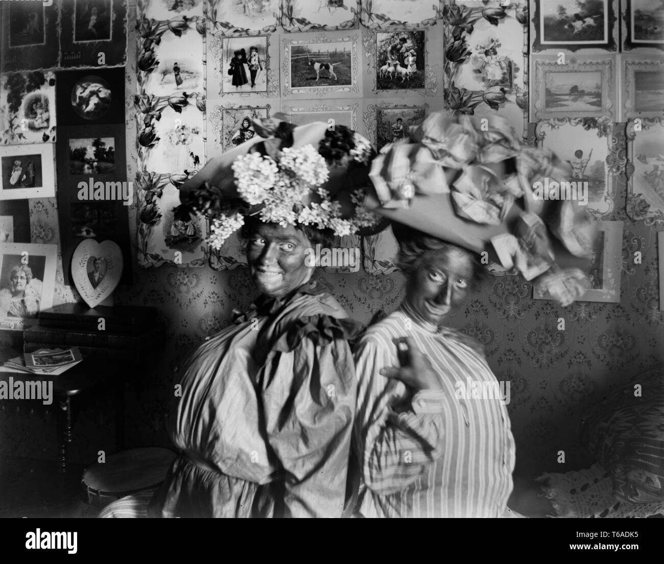 Two Victorian woman in blackface wearing large flowered hats playfully sit back to back, ca. 1900. Stock Photo