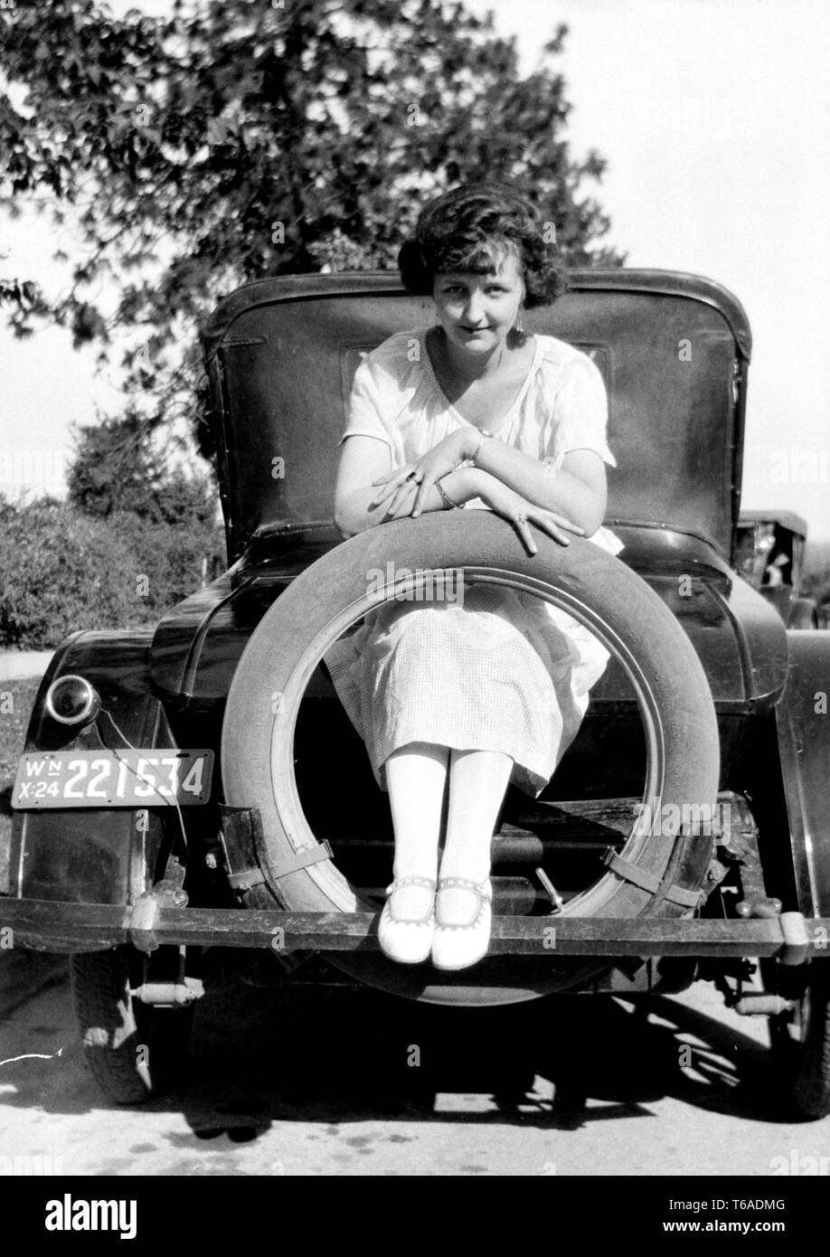 A young woman poses on the rear of a period car with her feet through the spare tire, ca. 1924. Stock Photo