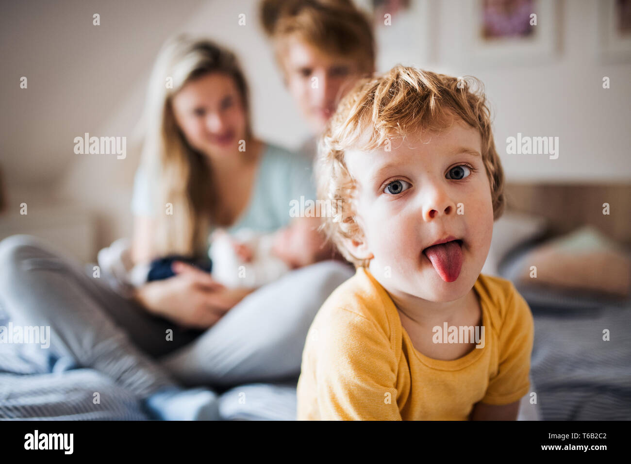 A small toddler boy with parents and newborn baby at home, sticking tongue out. Stock Photo