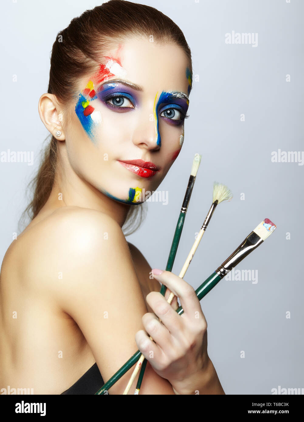 Young female painter with paint brush in hand and acrylic paint on face