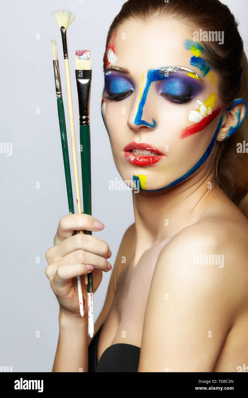 Young female painter with paint brush in hand and acrylic paint on face ...