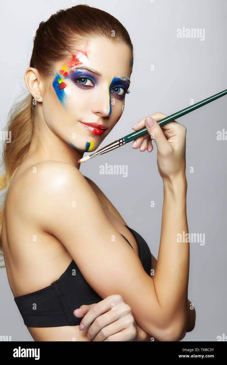 Young female painter with paint brush in hand and acrylic paint on face ...