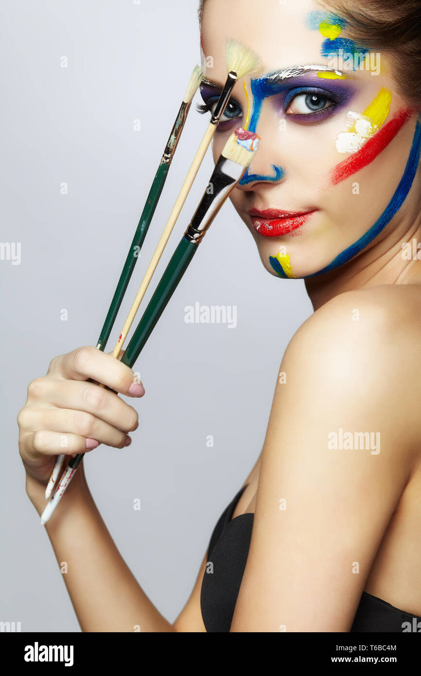 Young female painter with paint brush in hand and acrylic paint on face ...