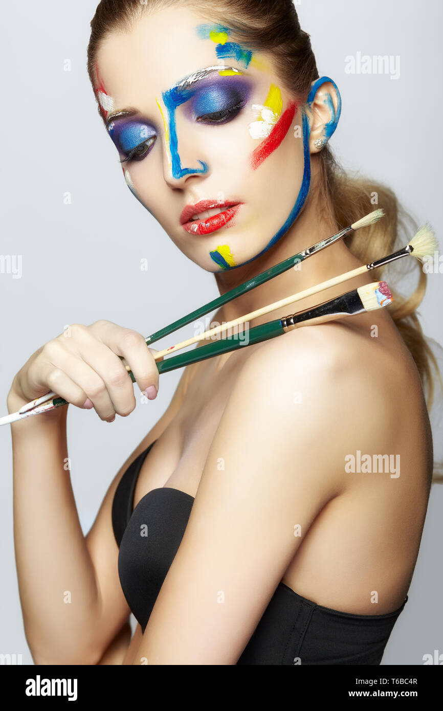 Young female painter with paint brush in hand and acrylic paint on face ...