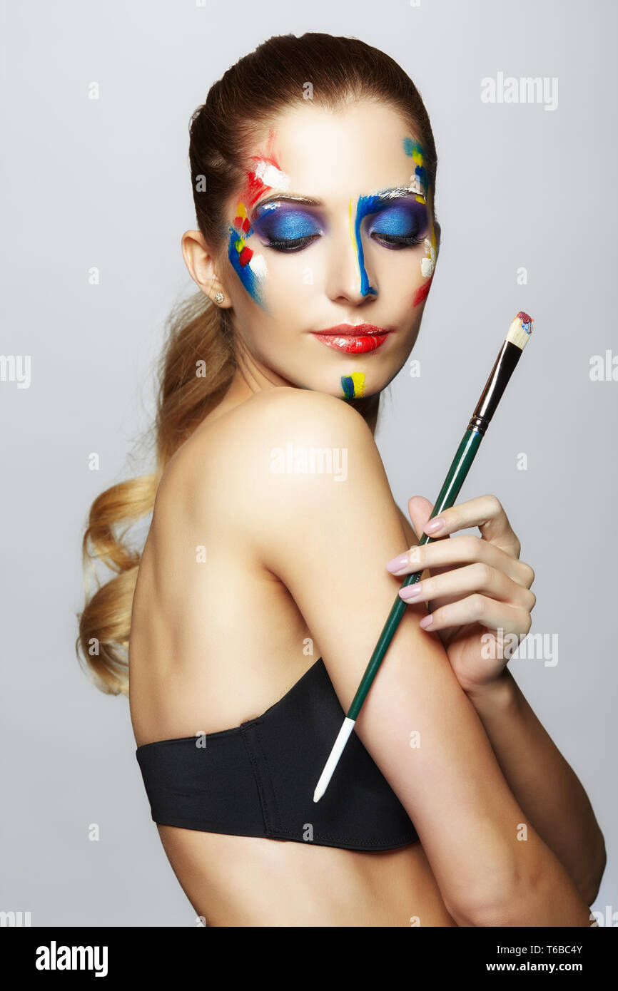 Young female painter with paint brush in hand and acrylic paint on face