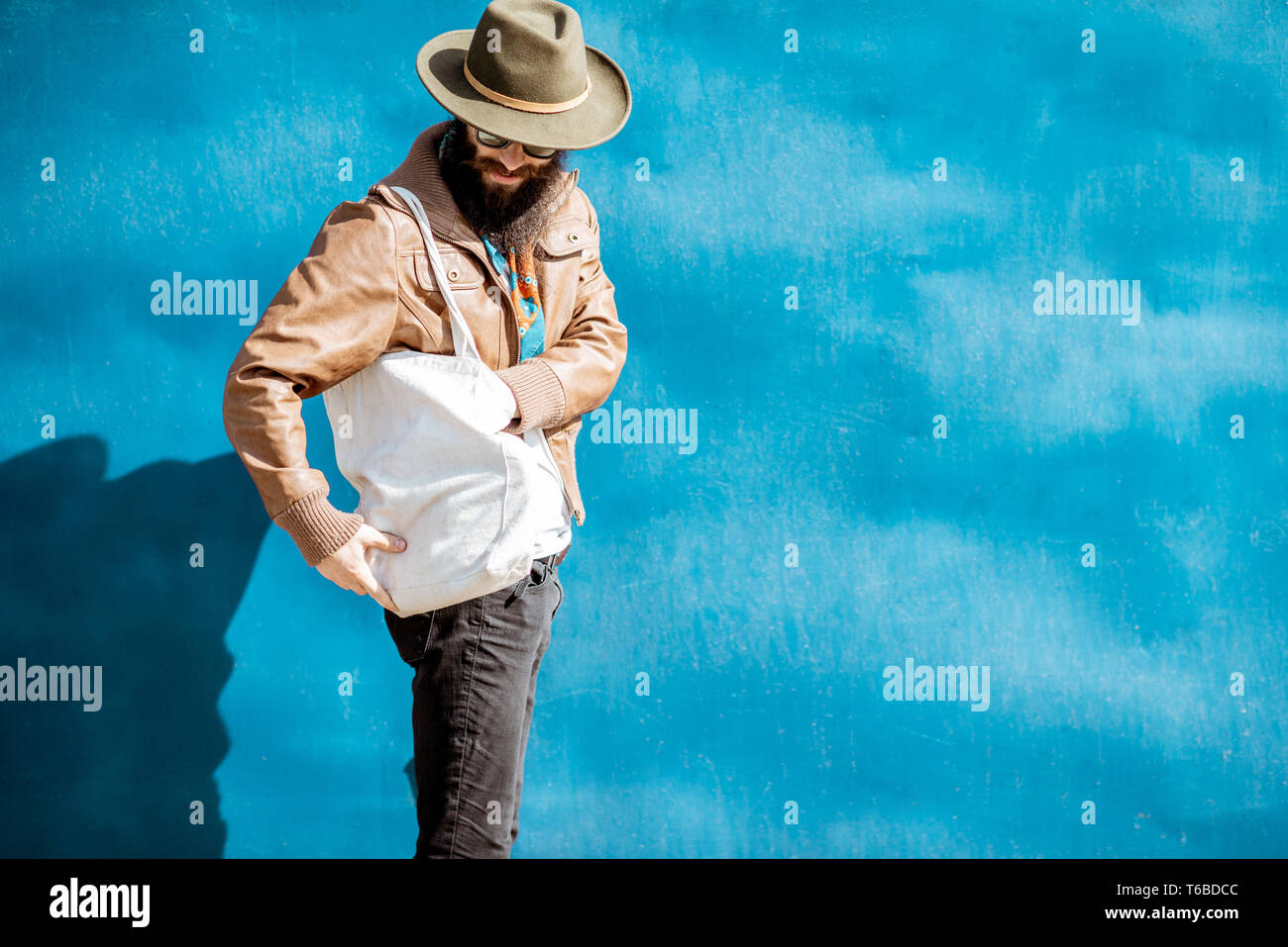 Portrait of a stylish bearded man dressed in jacket and hat standing with bag on the blue background outdoors Stock Photo