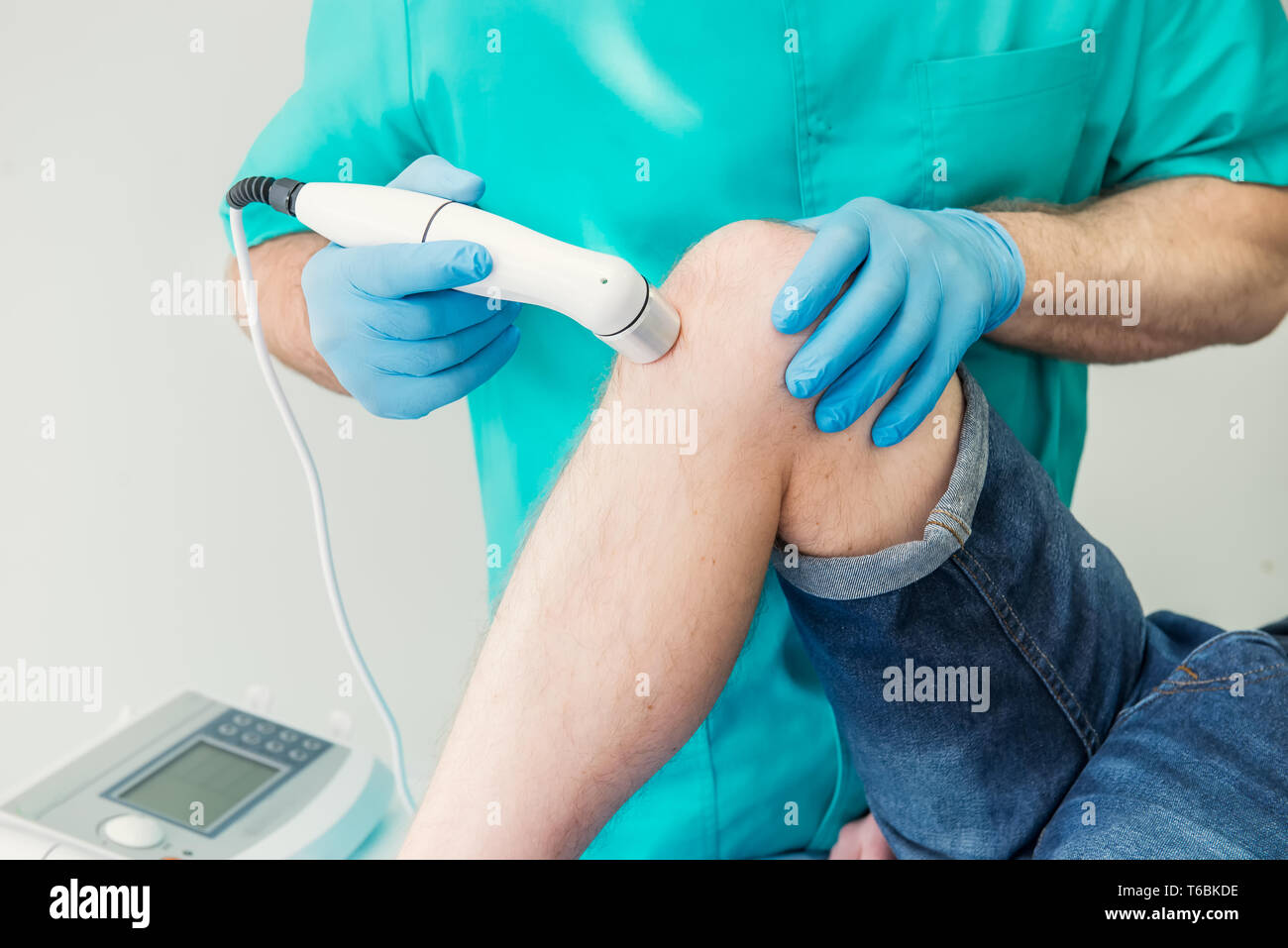 Close up Young man receiving laser or magnet therapy massage on a knee to less pain. A chiropractor treats patient's knee-joint in medical office. Neu Stock Photo
