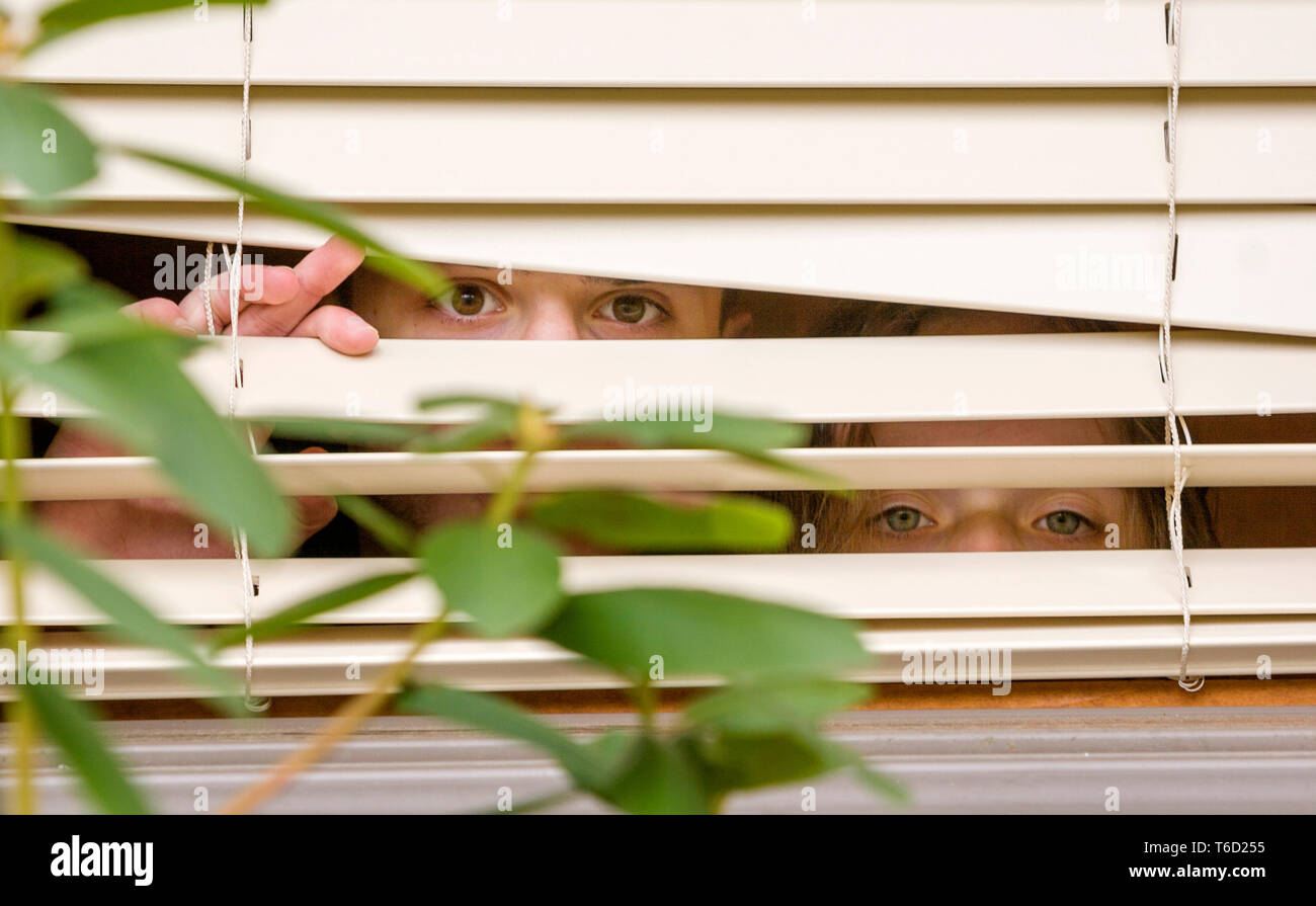 Brother and sister peeking through partially opened window blinds. Stock Photo