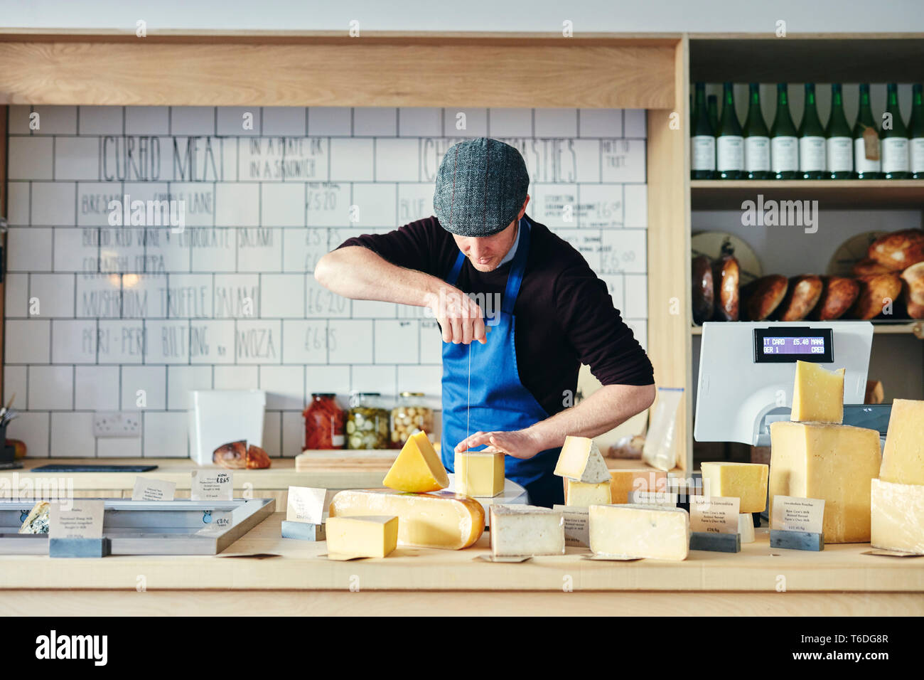 Cheesemonger cutting cheese with cheese wire surrounded by a variety of cheese on counter top Stock Photo