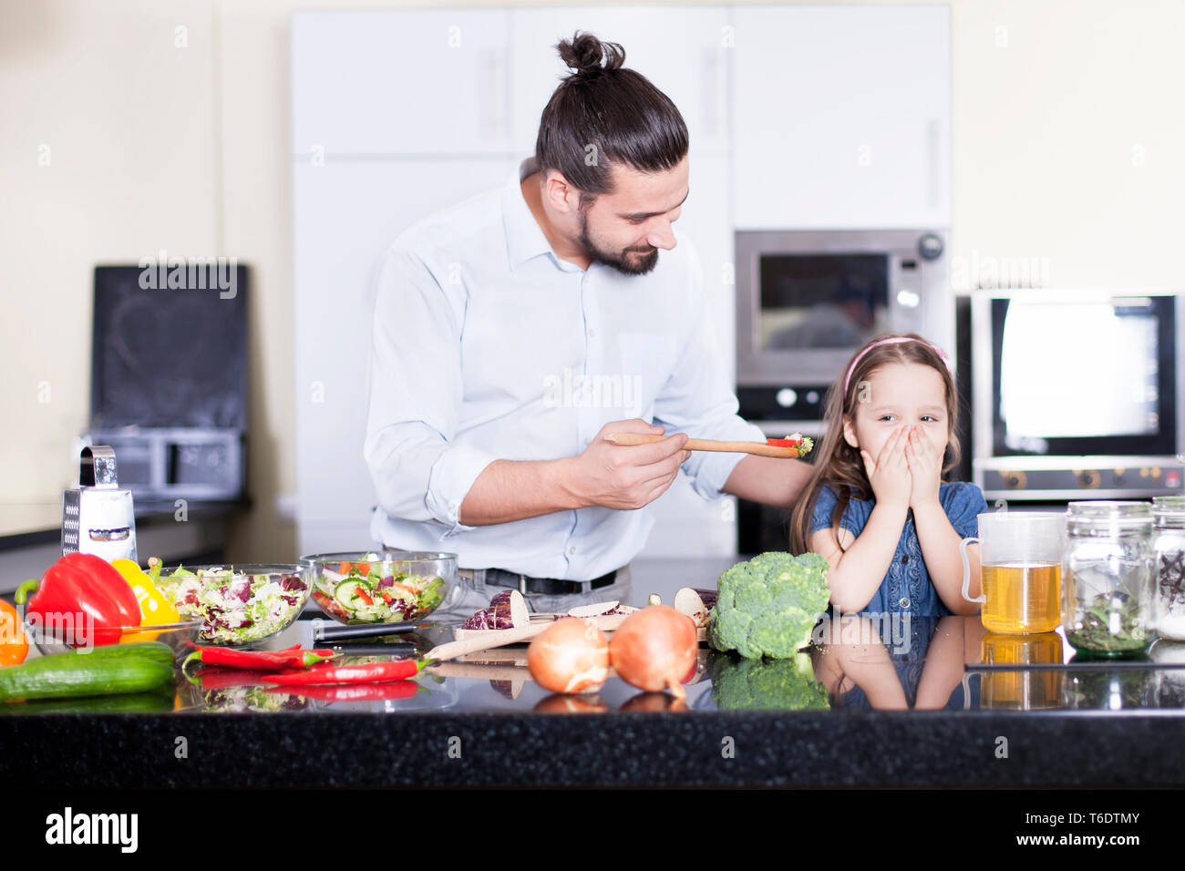Little girl refusing to eat a salad while cooking and having lunch together in the kitchen Stock Photo