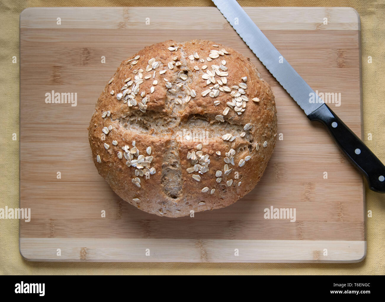 A round loaf of artisan whole-grain bread with scored crust, garnished with rolled oats, overhead shot, resting on a bamboo cutting board. Stock Photo