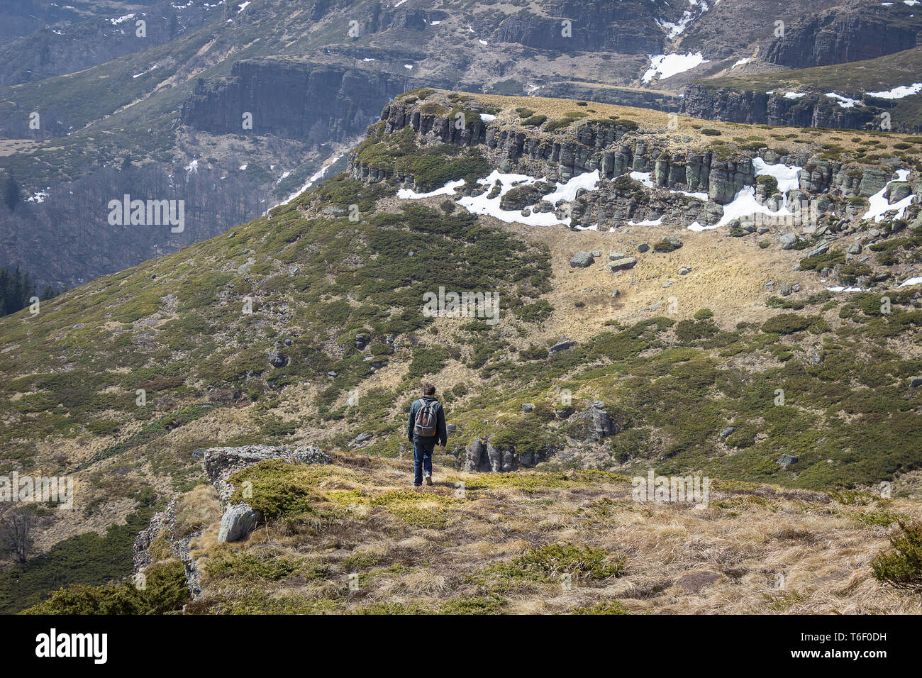 Mountain hiker walking down the rocky, sunlit highlands covered by snow, dry grass and juniper Stock Photo