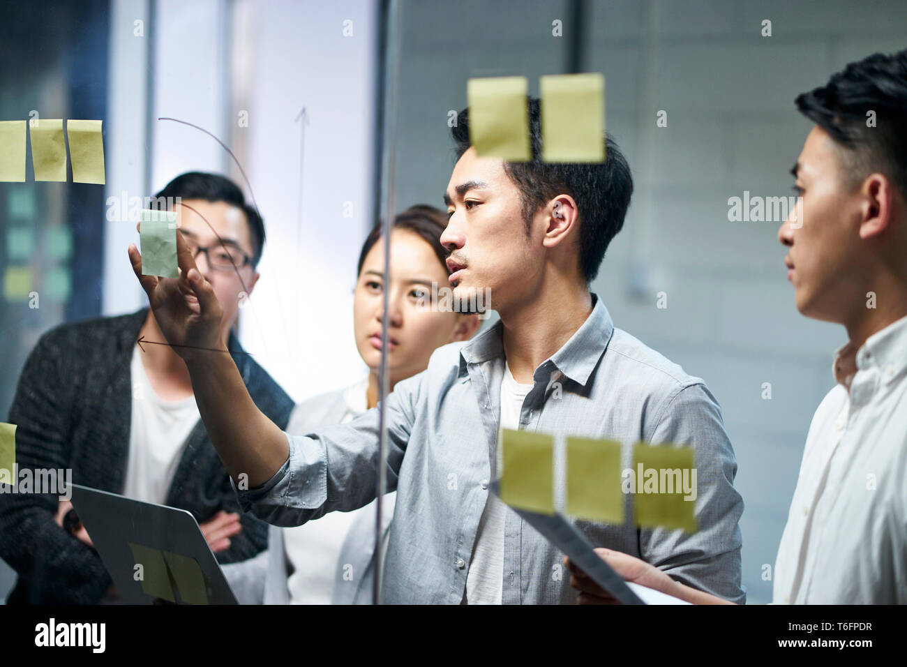 young asian entrepreneur of small company putting a adhesive note on glass in office during team meeting formulating business strategies. Stock Photo