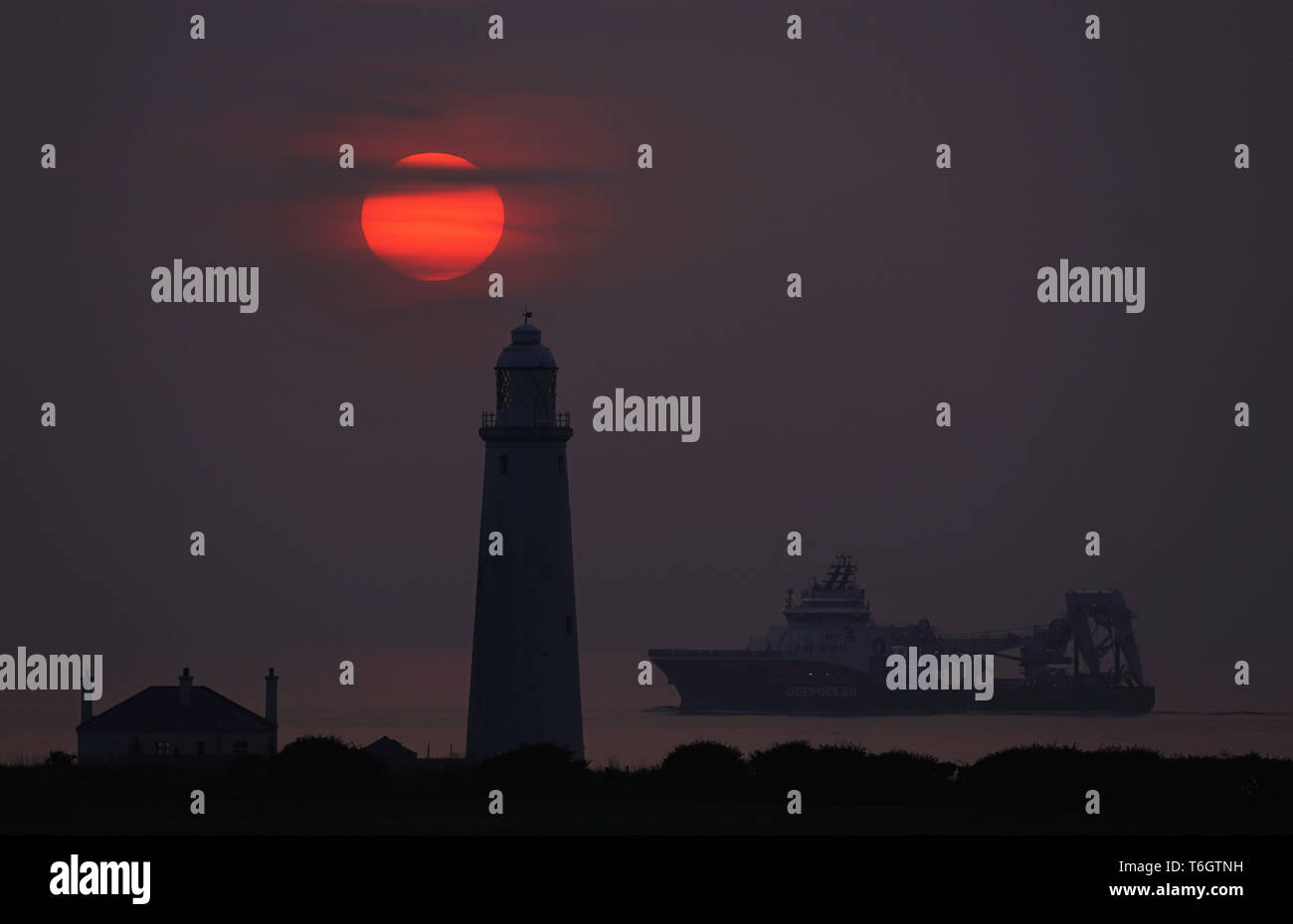 The sun rises through the mist at St Mary's lighthouse, Whitley Bay as the off shore supply ship Havila Phoenix heads into Blyth on the Northumberland coast. Stock Photo