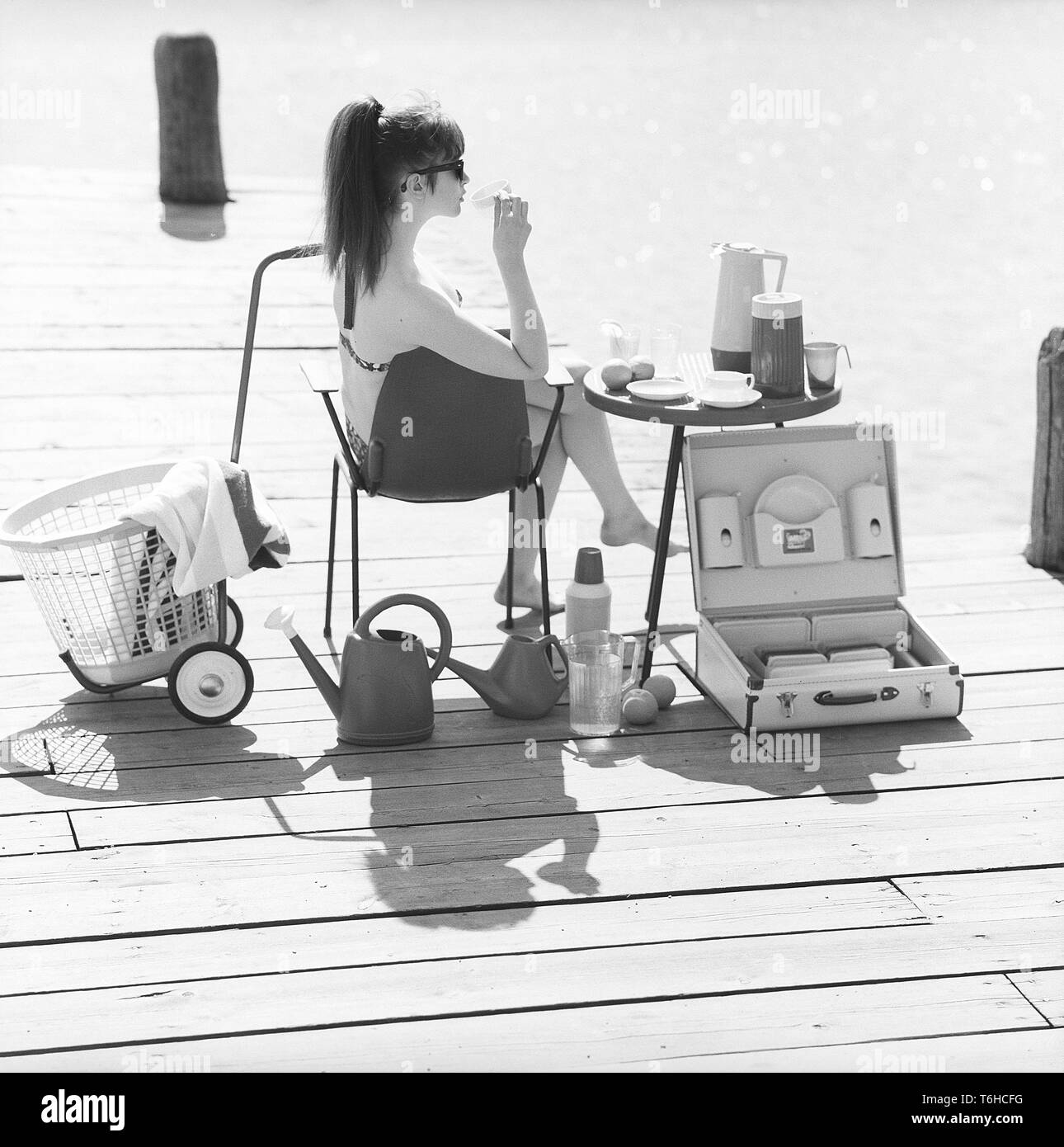1960s lifestyle. A young woman sitting in the sun wearing a bikini. In the picture there are many of the timely objects made of plastic that were popular at the time. Especially when being on a summer vacation or camping. A set of cups and saucers on the table. A practical case to transport them in. Sweden 1960s Photo Kristoffersson ref DB85-2 Stock Photo